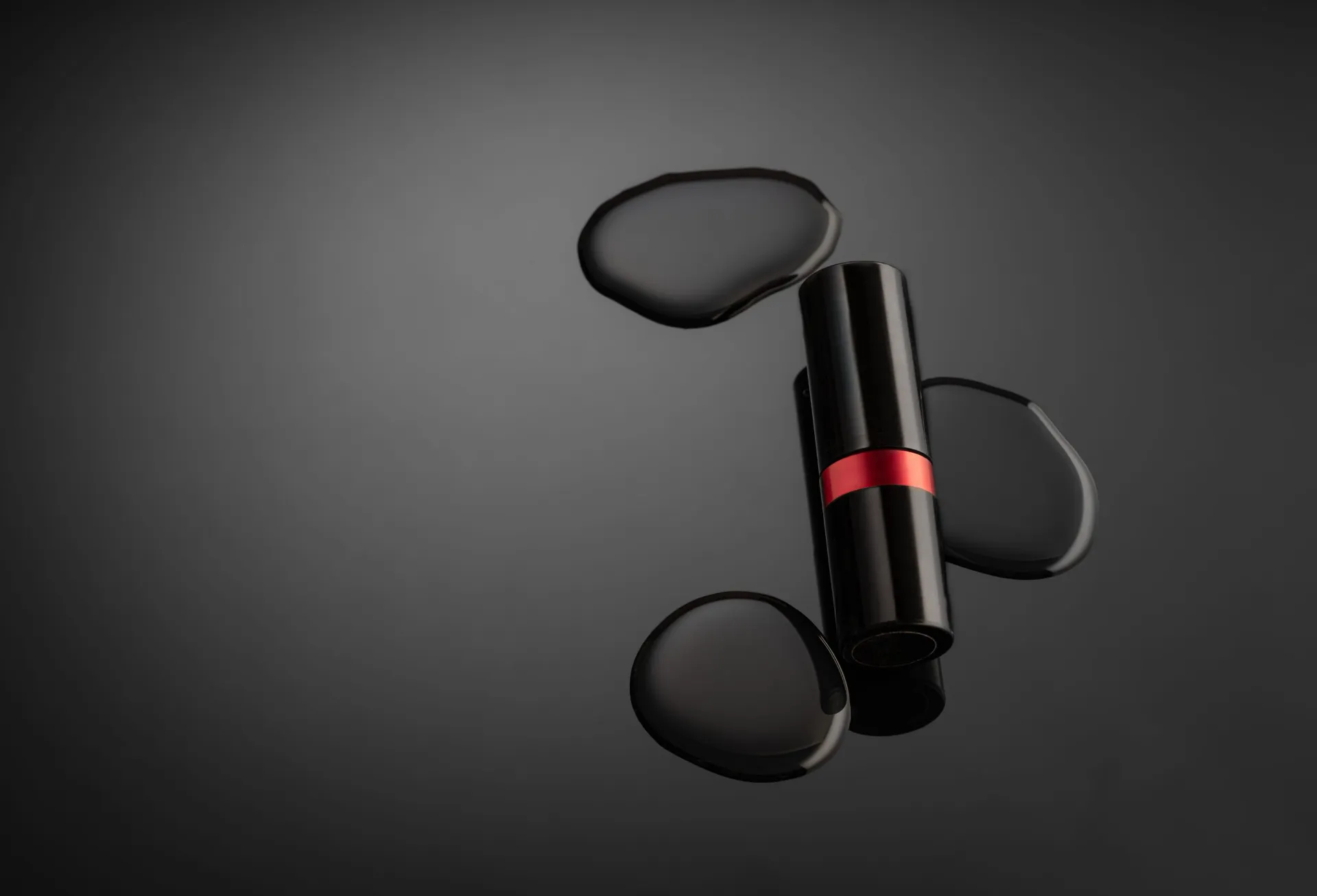 A black lipstick with a red band lying among glossy droplets on a dark grey surface.